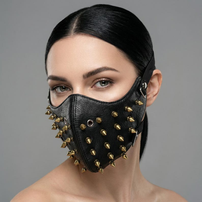 May include: Black leather face mask with gold-colored spikes. The mask has a contoured shape and adjustable straps. The spikes are evenly spaced across the mask's surface, adding a bold, edgy aesthetic. The mask is designed for a unique, alternative style.