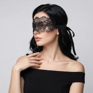 Puede incluir: Una máscara de ojos de encaje negro con un diseño floral. La máscara tiene un borde festoneado y está atada con cintas negras. La máscara es usada por una persona con cabello oscuro y un top negro con hombros descubiertos.