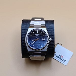 May include: A stainless steel Tissot PRX watch with a blue dial and silver hands. The watch is displayed on a black stand, with a white tag attached. The watch face has the Tissot logo and the text "PRX Powermatic 80".