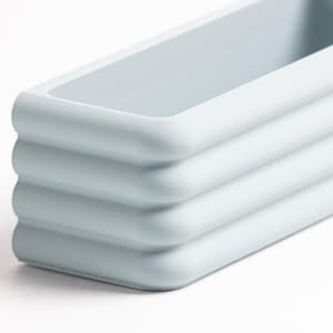 Puede incluir: Una bandeja rectangular de color azul claro con esquinas redondeadas y un dise&ntilde;o exterior acanalado y escalonado. El objeto presenta un acabado mate suave y un compartimento interior profundo y abierto, mostrando una forma estructurada.