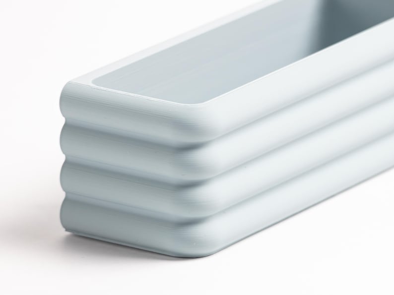 Puede incluir: Una bandeja rectangular de color azul claro con esquinas redondeadas y un dise&ntilde;o exterior acanalado y escalonado. El objeto presenta un acabado mate suave y un compartimento interior profundo y abierto, mostrando una forma estructurada.
