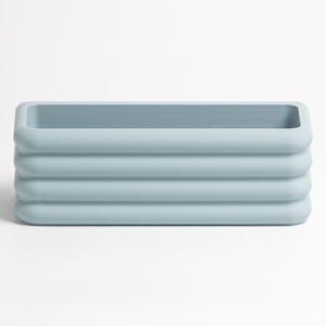 Puede incluir: Una maceta rectangular de color azul claro con un acabado mate suave y bordes redondeados. El exterior presenta tres nervaduras horizontales que envuelven todo el cuerpo, creando una apariencia apilada con una abertura central hueca.