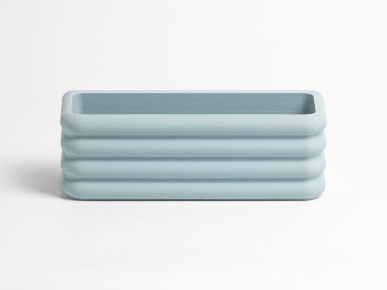 Puede incluir: Una maceta rectangular de color azul claro con un acabado mate suave y bordes redondeados. El exterior presenta tres nervaduras horizontales que envuelven todo el cuerpo, creando una apariencia apilada con una abertura central hueca.