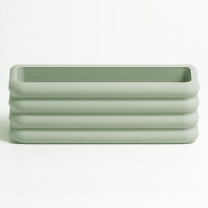 Puede incluir: Una maceta rectangular de color verde salvia con un acabado mate suave y cuatro crestas horizontales redondeadas a lo largo de las paredes exteriores. El recipiente presenta una parte superior abierta y un interior hueco.