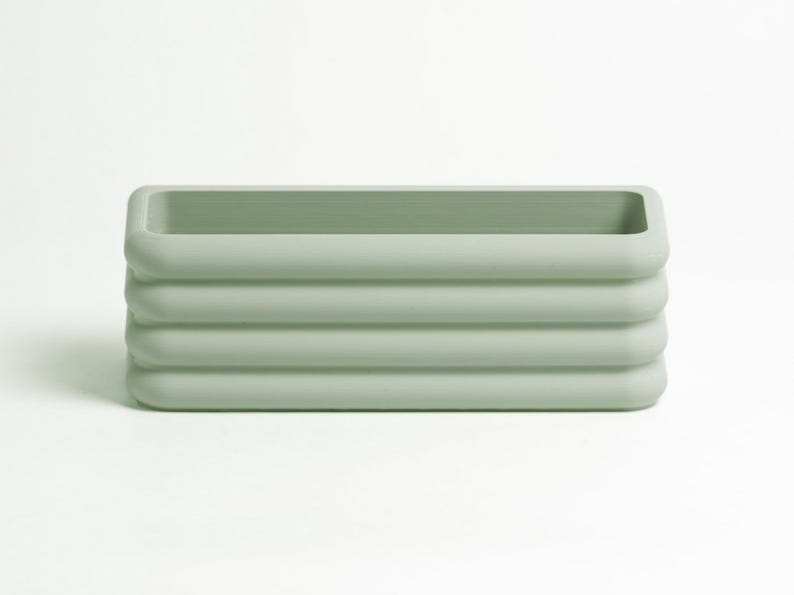 Puede incluir: Una maceta rectangular de color verde salvia con un acabado mate suave y cuatro crestas horizontales redondeadas a lo largo de las paredes exteriores. El recipiente presenta una parte superior abierta y un interior hueco.