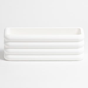 Puede incluir: Una maceta rectangular blanca con un acabado mate liso y una serie de cuatro crestas horizontales redondeadas a lo largo de los lados exteriores. El objeto presenta una parte superior abierta y un interior hueco, de aspecto uniforme.