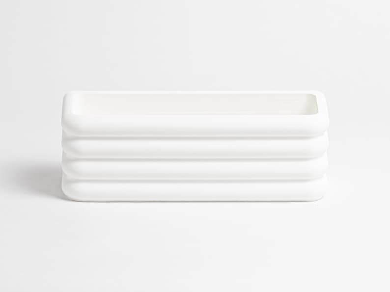 Puede incluir: Una maceta rectangular blanca con un acabado mate liso y una serie de cuatro crestas horizontales redondeadas a lo largo de los lados exteriores. El objeto presenta una parte superior abierta y un interior hueco, de aspecto uniforme.