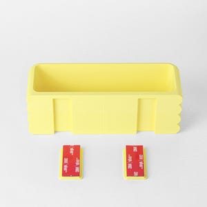 Puede incluir: Un contenedor de almacenamiento rectangular de pl&aacute;stico amarillo con un interior hueco y un dise&ntilde;o de borde festoneado en los lados. Dos peque&ntilde;os soportes de montaje rectangulares amarillos se colocan debajo del contenedor, cada uno con una tira adhesiva 3M roja.