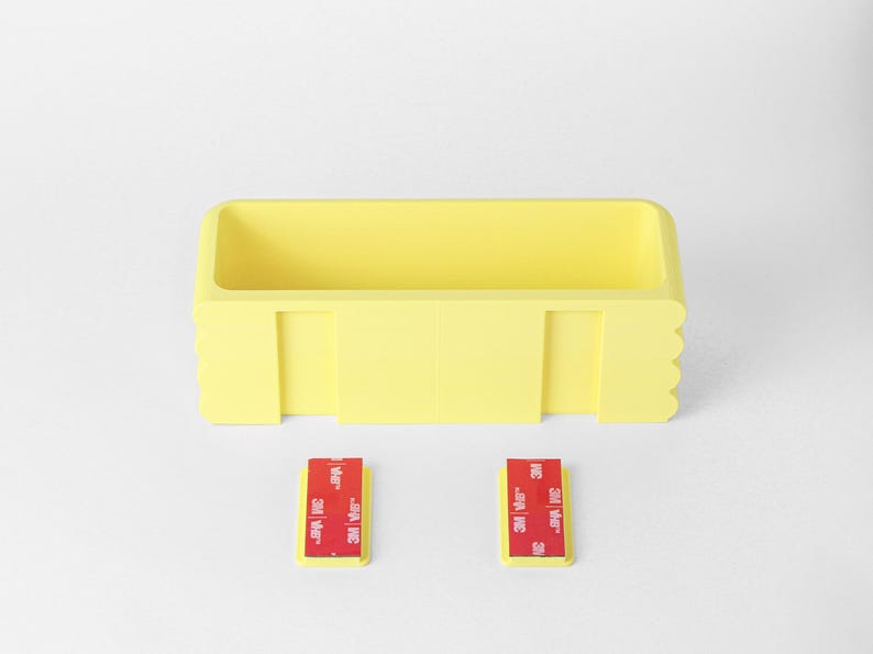Puede incluir: Un contenedor de almacenamiento rectangular de pl&aacute;stico amarillo con un interior hueco y un dise&ntilde;o de borde festoneado en los lados. Dos peque&ntilde;os soportes de montaje rectangulares amarillos se colocan debajo del contenedor, cada uno con una tira adhesiva 3M roja.