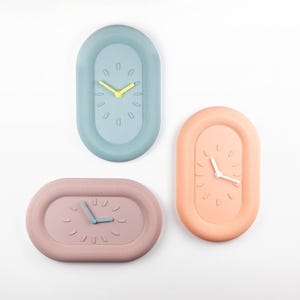 Könnte beinhalten: Drei ovale Wanduhren in Pastellfarben: Hellblau, Pfirsich und Mauve. Jede Uhr hat ein minimalistisches Design mit einem einfachen Zifferblatt und Zeigern. Die hellblaue Uhr hat gelbe Zeiger, die Pfirsichuhr hat weiße Zeiger und die mauvefarbene Uhr hat blaue Zeiger.