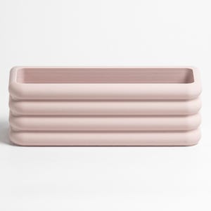 Puede incluir: Un recipiente rectangular de color rosa claro que presenta una serie de cuatro nervaduras horizontales redondeadas a lo largo de sus paredes exteriores. El objeto tiene un centro hueco y un acabado mate liso, con un aspecto limpio y uniforme.