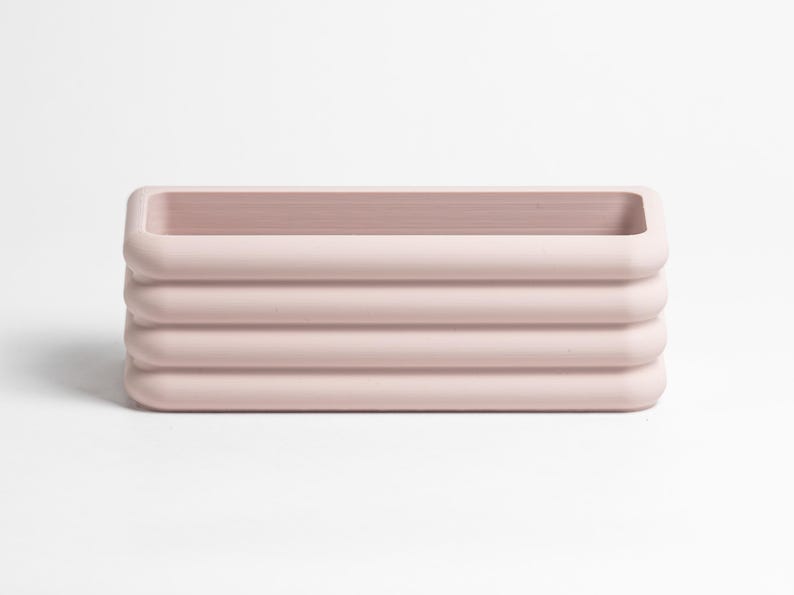 Puede incluir: Un recipiente rectangular de color rosa claro que presenta una serie de cuatro nervaduras horizontales redondeadas a lo largo de sus paredes exteriores. El objeto tiene un centro hueco y un acabado mate liso, con un aspecto limpio y uniforme.