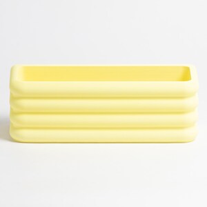 Puede incluir: Un recipiente rectangular de color amarillo p&aacute;lido que presenta una serie de cuatro nervaduras horizontales y redondeadas a lo largo de sus paredes exteriores. El objeto tiene un acabado mate liso y una parte superior abierta.