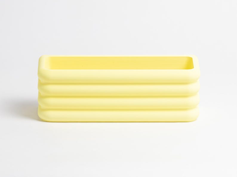Puede incluir: Un recipiente rectangular de color amarillo p&aacute;lido que presenta una serie de cuatro nervaduras horizontales y redondeadas a lo largo de sus paredes exteriores. El objeto tiene un acabado mate liso y una parte superior abierta.