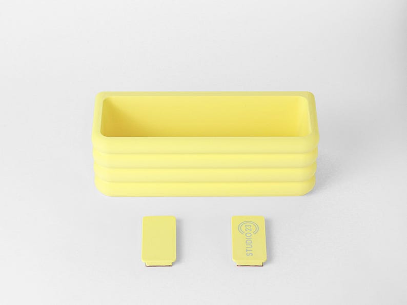 Puede incluir: Un recipiente rectangular amarillo con un dise&ntilde;o exterior acanalado. Debajo del recipiente hay dos peque&ntilde;os accesorios rectangulares amarillos, uno de los cuales tiene el texto "STUDIO 23" impreso en color gris claro en su superficie.