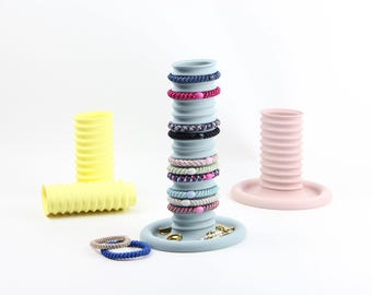 Puede incluir: Tres soportes cilíndricos acanalados en amarillo, azul claro y rosa pálido. El soporte azul claro sostiene una pila de gomas para el pelo de colores, con dos gomas adicionales en su base. El soporte amarillo está en posición horizontal.