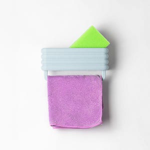 Może przedstawiać: Jasnoniebieski plastikowy uchwyt ścienny z poziomymi żebrowaniami. W g&oacute;rnej przegrodzie umieszczona jest jaskrawozielona prostokątna gąbka, a na dolnym drążku wisi złożona fioletowa ściereczka z mikrofibry.