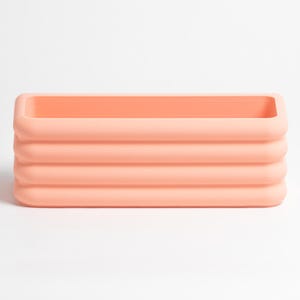 Puede incluir: Un recipiente rectangular de color melocot&oacute;n con un centro hueco y esquinas redondeadas. El exterior presenta cuatro bandas horizontales acanaladas y apiladas que crean una apariencia texturizada y en capas en los lados del objeto.
