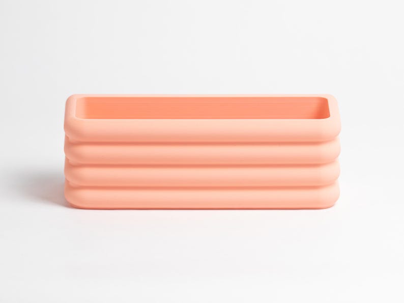 Puede incluir: Un recipiente rectangular de color melocot&oacute;n con un centro hueco y esquinas redondeadas. El exterior presenta cuatro bandas horizontales acanaladas y apiladas que crean una apariencia texturizada y en capas en los lados del objeto.