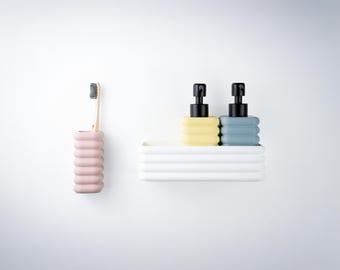 Puede incluir: Un juego de baño que incluye un portacepillos de dientes rosa claro acanalado con un cepillo de madera, y una bandeja rectangular blanca acanalada con dos dispensadores de jabón, uno amarillo pálido y otro azul grisáceo, con bombas negras.
