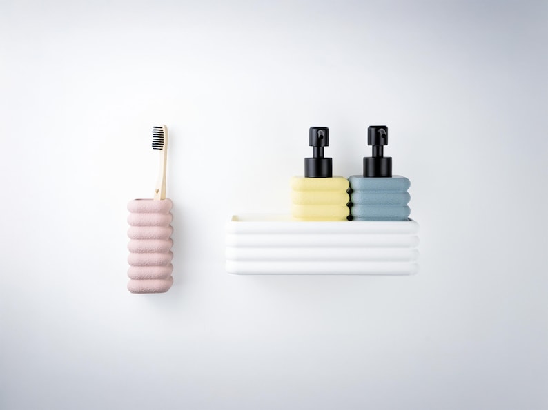 Puede incluir: Un juego de ba&ntilde;o que incluye un portacepillos de dientes rosa claro acanalado con un cepillo de madera, y una bandeja rectangular blanca acanalada con dos dispensadores de jab&oacute;n, uno amarillo p&aacute;lido y otro azul gris&aacute;ceo, con bombas negras.