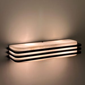 Könnte beinhalten: Rechteckige Wandleuchte mit geschichtetem Design. Die Leuchte hat eine weiße, gebogene Front mit drei horizontalen schwarzen Streifen. Das Licht strahlt ein warmes, einladendes Licht aus.