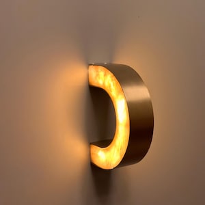 May include: A modern, illuminated wall sconce in the shape of a letter "C". The fixture is made of a brushed gold metal with a warm, amber-colored light emanating from within. The sconce is mounted on a neutral-colored wall.