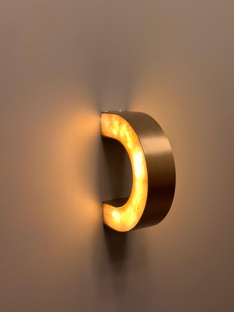 May include: A modern, illuminated wall sconce in the shape of a letter "C". The fixture is made of a brushed gold metal with a warm, amber-colored light emanating from within. The sconce is mounted on a neutral-colored wall.