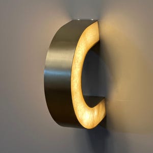 May include: A modern, wall-mounted light fixture in a curved, C-shape design. The fixture has a brushed metal exterior and an illuminated, warm-toned interior. The light is mounted on a gray wall, creating a contemporary aesthetic.