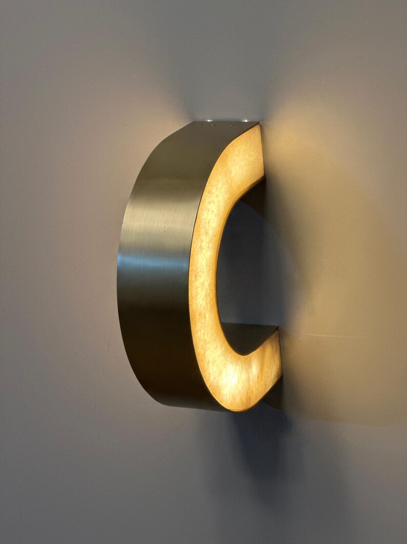 May include: A modern, wall-mounted light fixture in a curved, C-shape design. The fixture has a brushed metal exterior and an illuminated, warm-toned interior. The light is mounted on a gray wall, creating a contemporary aesthetic.