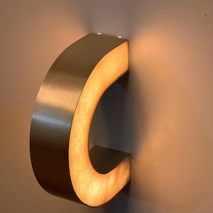 May include: A brushed metal letter "C" mounted on a wall. The interior of the letter is illuminated with a warm, soft light. The letter is a decorative element, possibly for signage or interior design.