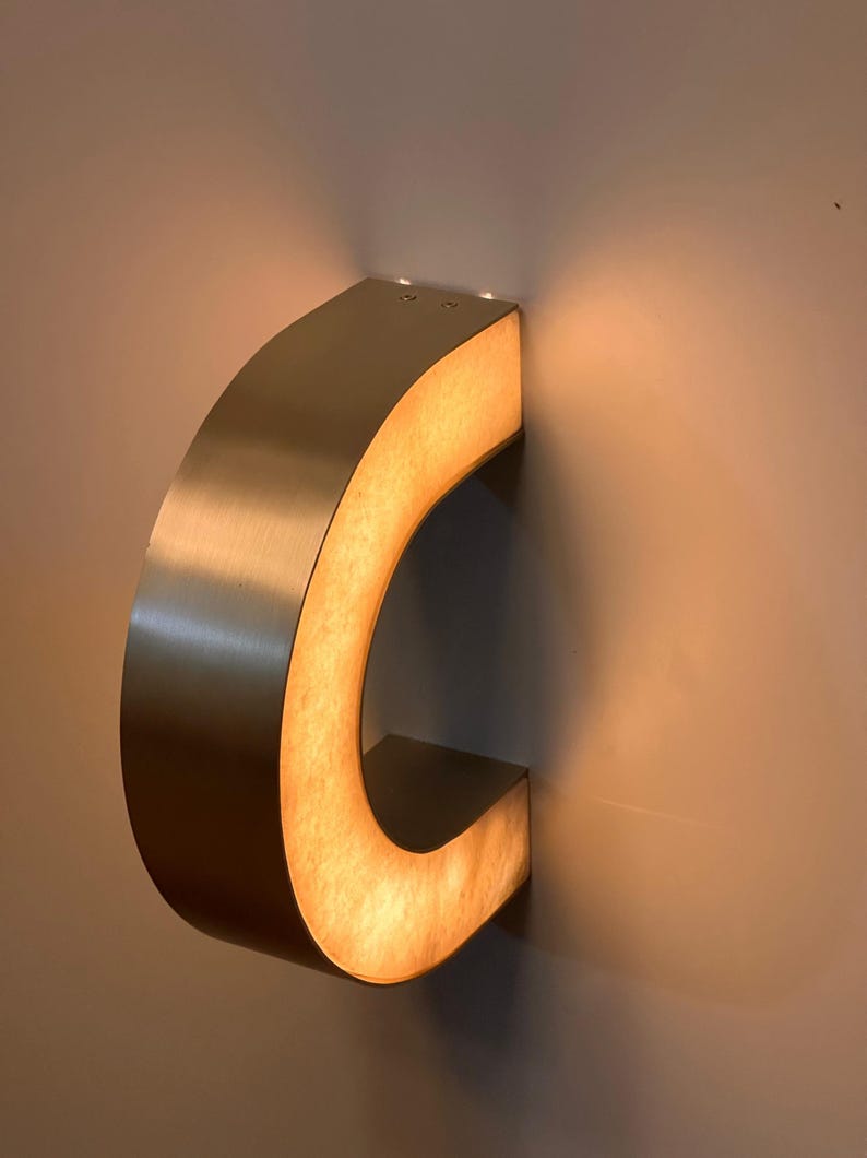 May include: A brushed metal letter "C" mounted on a wall. The interior of the letter is illuminated with a warm, soft light. The letter is a decorative element, possibly for signage or interior design.
