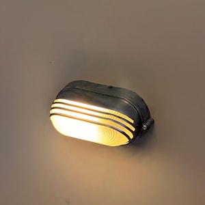Op de afbeelding: Een ovaalvormige, antieke wandlamp. De lamp heeft een donkere, metalen afwerking met horizontale lijnen en een helder, warm licht dat uit het matglas komt. De lamp is bevestigd aan een neutraal gekleurde muur.