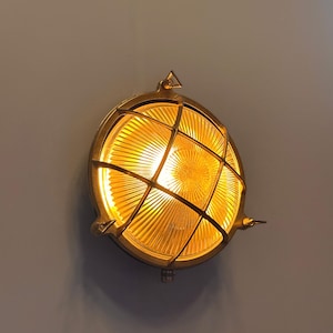 May include: A round, brass-colored wall-mounted light fixture with a grid-like cover. The light emits a warm, yellow glow. The fixture is mounted on a gray wall, creating a contrast between the light and the background. The design is reminiscent of nautical or industrial styles.