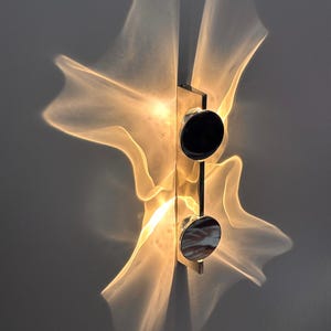 Può includere: Una lampada da parete con una barra metallica verticale e due accenti circolari neri. La luce emette un motivo luminoso caldo, traslucido e ondulato che si estende verso l'esterno dal centro, creando un effetto morbido e diffuso.
