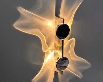 May include: A wall-mounted light fitting featuring a vertical metallic bar with two circular black accents. The light emits a warm, glowing, translucent, and wavy light pattern that extends outwards from the centre, creating a soft, diffused effect.
