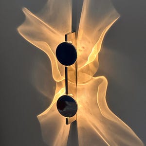 Könnte beinhalten: Moderne Wandleuchte mit minimalistischem Design. Sie verfügt über einen rechteckigen Metallrahmen mit zwei kreisförmigen, dunkel gefärbten Akzenten. Das Licht erzeugt ein einzigartiges, abstraktes Muster an der Wand mit weichen, fließenden Formen und warmen Tönen.