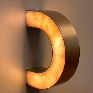 May include: A modern, illuminated wall sconce in the shape of a letter "C". The fixture has a warm, amber glow emanating from within, framed by a metallic, curved exterior. The sconce is mounted on a neutral-colored wall.