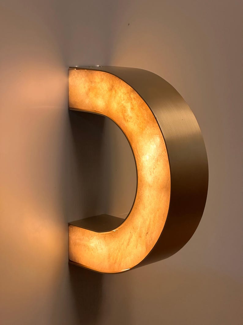 May include: A modern, illuminated wall sconce in the shape of a letter "C". The fixture has a warm, amber glow emanating from within, framed by a metallic, curved exterior. The sconce is mounted on a neutral-colored wall.