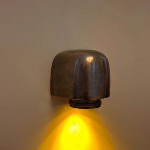 Può includere: Una lampada da parete in metallo con finitura scura spazzolata e un alloggiamento arrotondato a forma di cupola. L'apparecchio presenta una base circolare con un bordo testurizzato ed emette una luce gialla calda e brillante verso il basso.