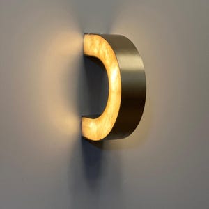 May include: A modern, curved wall sconce with a warm, glowing interior. The sconce has a metallic, bronze-colored exterior and is mounted on a gray wall. The light source is concealed within the curved design, creating a soft, ambient glow.