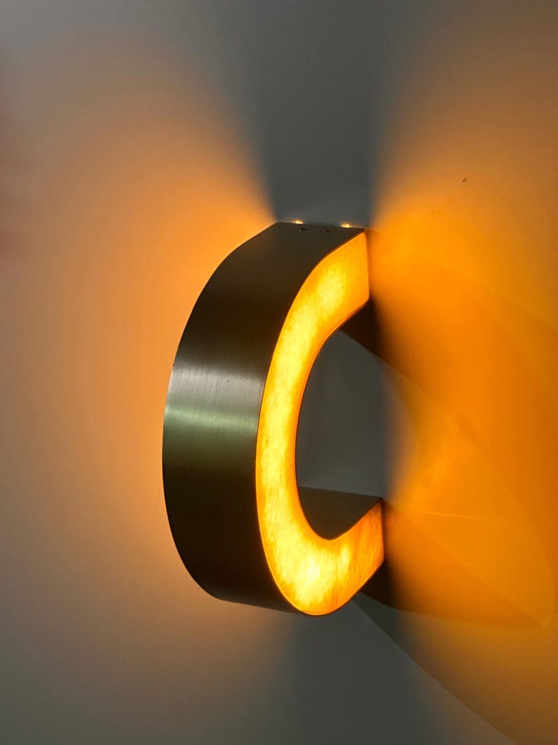 May include: A metallic letter "C" illuminated with warm, yellow light. The letter is mounted on a wall, casting shadows. The lighting creates a glowing effect, highlighting the curved shape of the letter.