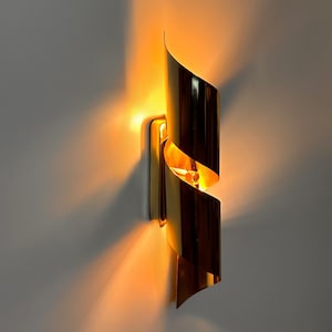 Könnte beinhalten: Goldfarbene Wandleuchte mit bandartigem Design. Die Leuchte ist an einer Wand montiert und strahlt ein warmes, einladendes Licht aus. Das Design weist eine geschwungene, geschichtete Form auf und erzeugt eine moderne Ästhetik.