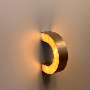 May include: A modern, C-shaped wall sconce with a warm, golden glow. The fixture is made of a brushed metal material and is mounted on a neutral-colored wall. The light source is hidden, creating a soft, ambient illumination.