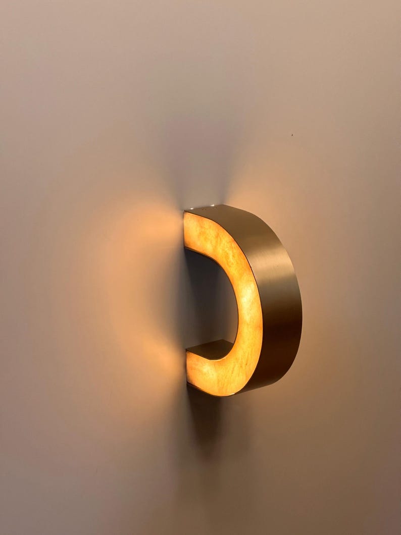 May include: A modern, C-shaped wall sconce with a warm, golden glow. The fixture is made of a brushed metal material and is mounted on a neutral-colored wall. The light source is hidden, creating a soft, ambient illumination.