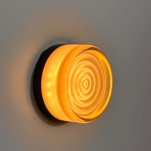 May include: A wall-mounted light fixture with a circular, amber-colored glass shade. The shade features a concentric spiral design and is encased in a dark, circular frame. The light emits a warm glow, suggesting an interior lighting product.