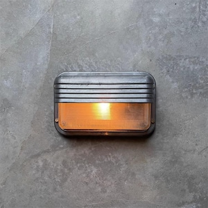 May include: A rectangular wall light with a brushed metal finish, mounted on a grey concrete wall. The light has horizontal ridges across the top and an amber-coloured lens. The light is on, emitting a warm glow. The fixture is industrial in style.