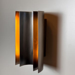 May include: A contemporary wall light with a brushed metal finish. The design incorporates vertical, curved panels, creating a layered effect. Warm light shines from the gaps between the panels, producing a soft glow. This light fixture is for indoor use.