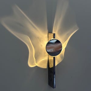 Könnte beinhalten: Eine Wandleuchte mit einem vertikalen, rechteckigen Chromsockel und einer zentralen, kreisförmigen Chromscheibe. Die Lampe strahlt ein warmes, diffuses Licht aus, das weiche, flügelartige Lichtmuster auf die Wandfläche wirft.