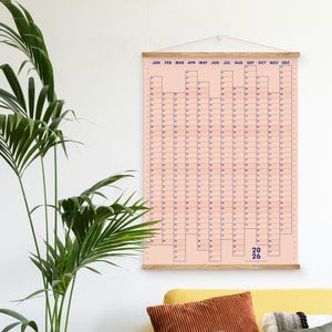 Peut inclure: Un calendrier annuel rose pour 2026 avec des lignes et du texte bleus, suspendu à une tringle en bois. Un canapé jaune avec des coussins marron et verts est en dessous. Un pot de plante dans un panier tressé est à gauche.